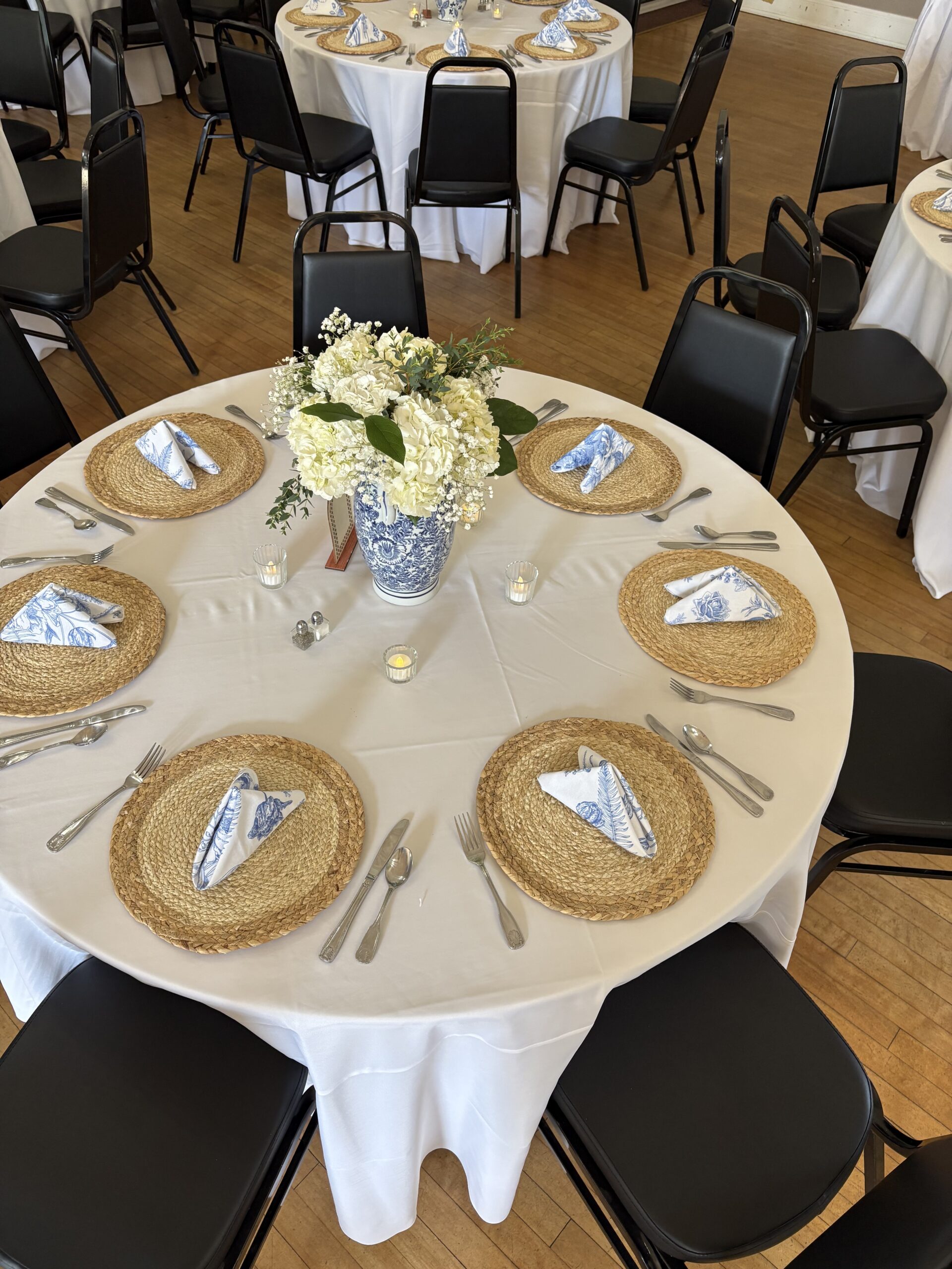 A round table with white linens, woven placemats, blue-and-white napkins, and a floral centerpiece.